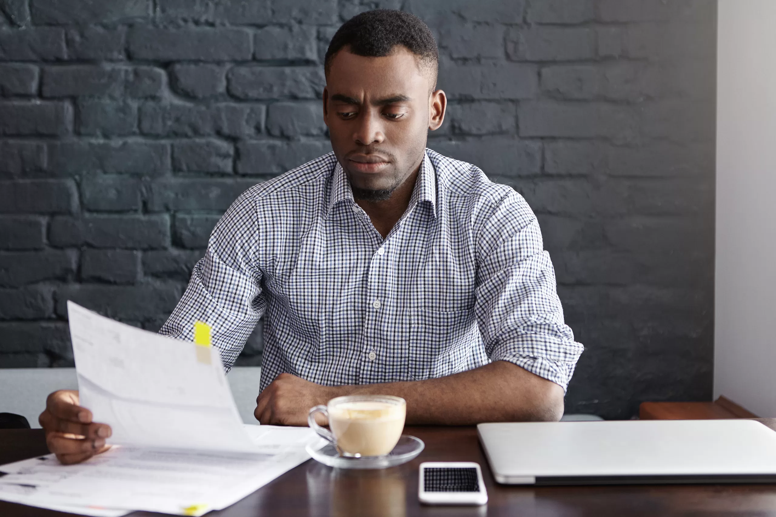 Indoor shot of serious African-American financier studying financial papers during coffee break at cafe, reading information with concentrated or worried look, siting against brick wall background