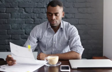 Indoor shot of serious African-American financier studying financial papers during coffee break at cafe, reading information with concentrated or worried look, siting against brick wall background